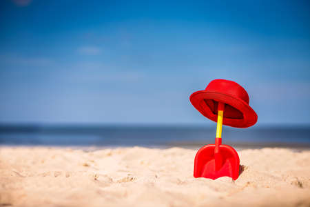 Red and yellow plastic childrens toy shovel and red straw hat on a beautiful white sand seashoreの写真素材