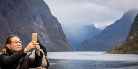 Flam, Norway -  October 2017 :  Chinese or japanese tourist taking pictures of beautiful scenery on his mobile during Norway in a nutshell sightseeing tour through the magnificent Aurlandsfjord and  Naeroyfjord fiordsのeditorial素材