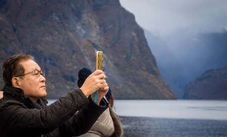 Flam, Norway -  October 2017 :  Chinese or japanese tourist taking pictures of beautiful scenery on his mobile during Norway in a nutshell sightseeing tour through the magnificent Aurlandsfjord and  Naeroyfjord fiordsのeditorial素材
