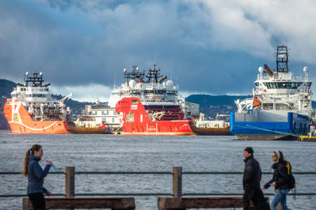 Bergen, Norway -  October 2017 : People walking on a waterfront promenade with large cargo ships in...