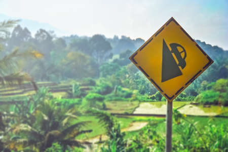 Yellow road sign warning of steep gradient of roads in Sumatran rural countryside, Indonesiaの写真素材