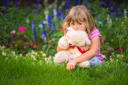 Adorable toddler girl hugging her favourite teddy bear in summer park on beautiful sunny day with yellow flowers in the backgroundの写真素材