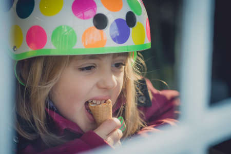 Young girl wearing bicycle helmet sitting in a cafe and eating ice cream in a cone