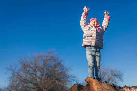 Cute little girl shouting from joy while standing on the top of large cut tree log in the park in springの写真素材