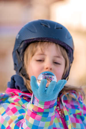 Winter portrait of a little girl wearing ski helmet and holding small glass ball with her own reflectionの写真素材