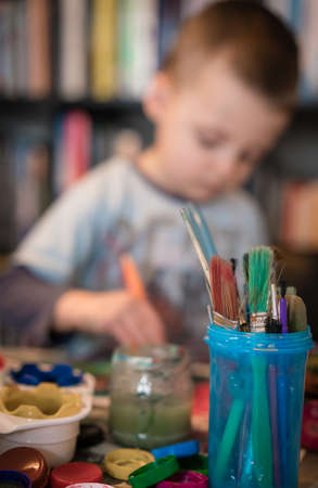 Little Caucasian boy sitting at a table at home and having fun with kids paint