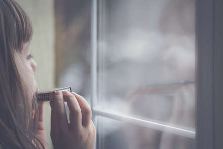 Sad Caucasian girl standing by the window and playing harmonica at home