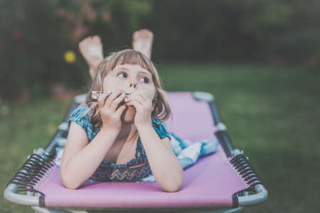 Barefoot girl dressed in blue dress lying on the reclining garden bed and playing on her small...