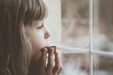 Sad Caucasian girl standing by the window and playing harmonica at home