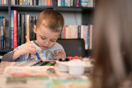 Little caucasian boy sitting at a table at home and having fun with kids paint