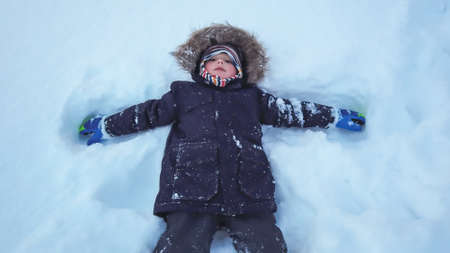 Little boy lying in a deep snow after heavy snowfall, moving his arms and legs to clear snow around...
