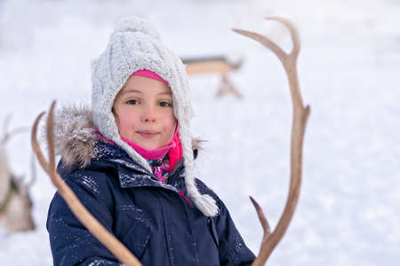 Portrait of a cute young girl in a warm winter jacket feeding reindeer in the winter, Tromso region,...