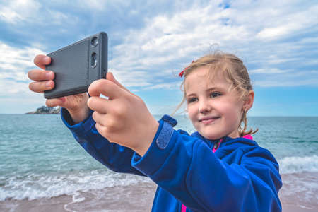 Cute little blond Caucasian girl taking selfie with mobile phone on the beach in autumn