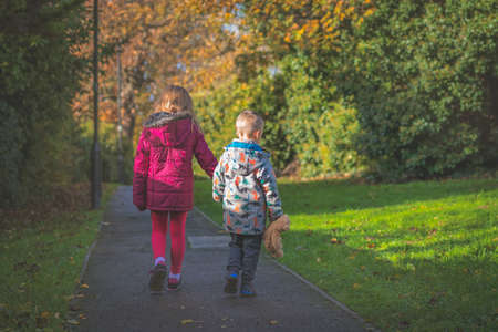 Boy holding hs favourite teddy bear soft toy holding hands with his sister and walking together on a path in the countryside in autumnの写真素材