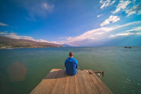 Male traveller sitting on a wooden pier and admiring the beauty of Ohrid Lake, Northern Macedonia