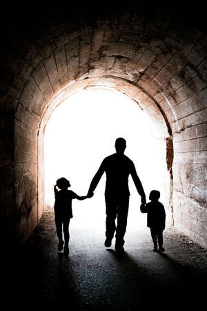 Father walking with his two little siblings children, boy and girl, towards the light at the end of a long dark tunnelの写真素材
