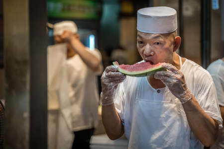 Xian, China -  July 2019 : Chinese chef taking his worki break and eating delicious and ripe slice of watermelon on the street in the Muslim Quarterのeditorial素材