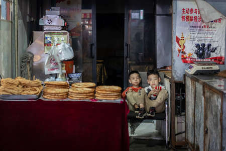 Xian, China -  August 2019 :  Two little boys playing games on a small mobile device while sitting on chairs in a small local bakery on the street in Muslim Quarterのeditorial素材