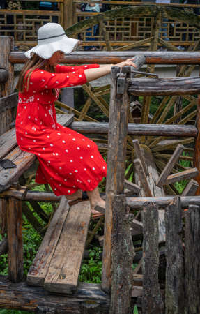 Wullingyuan, China -  August 2019 : Female dressed in a red dress using her legs to pull levers on a wheel of an old Water wheel in Huanglong Yellow Dragon Cave scenic area, Hunan provinceのeditorial素材