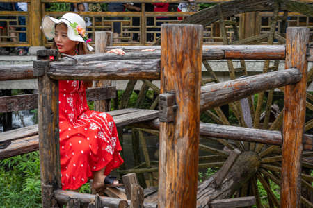 Wullingyuan, China -  August 2019 : Female dressed in a red dress using her legs to pull levers on a wheel of an old Water wheel in Huanglong Yellow Dragon Cave scenic area, Hunan provinceのeditorial素材