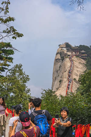 Huashan, China -  August 2019 : Chinese woman taking selfie picture on her phone with the crowds of tourists walking on a narrow ridge on a mountain trail to the West Peak in the background, Huashan mountain, Shaanxi Provinceのeditorial素材
