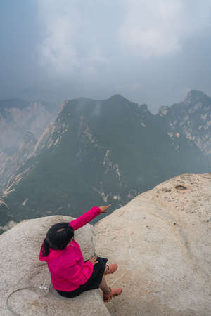 Huashan, China -  August 2019 : Tired chinese woman with sitting on a rock and pointing to the stunning landscape visible from the summit of the West Peak on Huashan mountain, Xian, Shaanxi Provinceのeditorial素材