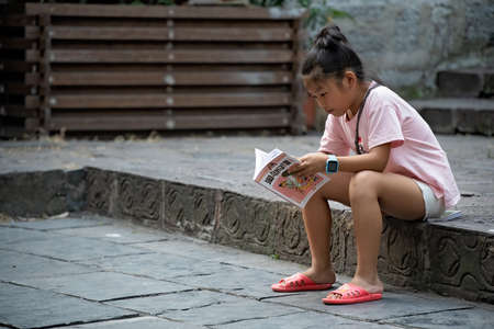 Feng Huang, China -  August 2019 : Young chinese girl sitting on a pavement and reading childrens book, Feng Huang Old Townのeditorial素材
