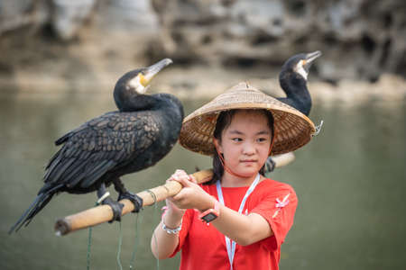 Guilin, China -  August 2019 : Cute little chinese girl holding bamboo stick with cormorant birds and posing for souvenir photo in front of the landmark Elephant Trunk Hill Arch and the Li Lijiang River, Guangxi Provinceのeditorial素材