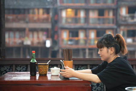 Feng Huang, China -  August 2019 : Chinese woman sitting in a cafe restaurant and using her tablet to watch clips or work, Fenghuang Old Townのeditorial素材