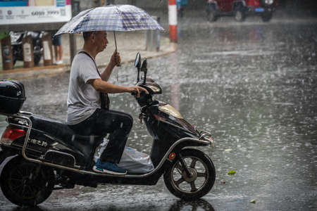 Chengdu, China - July  2019 : Chinese man riding on a scooter and holding umbrella  during tropical monsoon rain in Chengdu, Sichuan provinceのeditorial素材