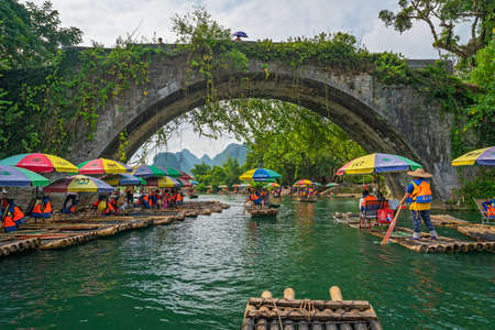 Yangshuo, China -  August 2019 : Small local tourist passenger bamboo boats carrying people under the historic Yulong Qiao bridge on the trip along the scenic Yulong river in Yangshuoのeditorial素材
