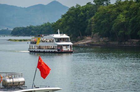 Guilin, China - August 2019 : Red Chinese national  flag fluttering on a mast on the deck of sightseeing boatt full of tourists departing on a trip on the magnificent Li river from Guilin to Yangshuoのeditorial素材