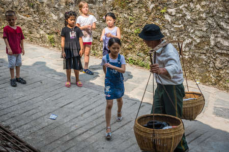 Pingan, China -  August 2019 : Curious village children surrounding Old Chinese man carrying heavy load of fruits for sale in Pingan, Longji Rice Terraces, Heping District, Longsheng County, Guilin Prefectureのeditorial素材