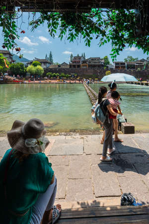 Feng Huang, China -  August 2019 : Older woman resting in tree shadow and watching people crossing waters on stepping stones on Tuojiang river, flowing through the centre of Fenghuang Old Townのeditorial素材