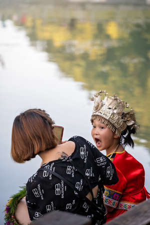 Feng Huang, China -  August 2019 : Mother taking picture of her beautiful young Chinese little girl daughter dressed in traditional folk costume and sitting on river shore in Fenghuang, Hunan Provinceのeditorial素材