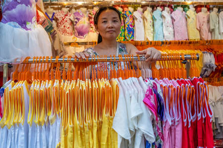 Chongqing, China - August 2019 : Chinese lady standing behind the rack with ladies dresses for sale in textile shop in Ci Qi Kou Ancient townのeditorial素材