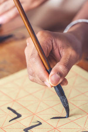 A closeup shot of a hand holding a brush with ink doing calligraphy on a sheet of paper