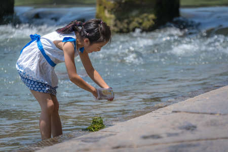 Feng Huang, China -  August 2019 : Cute little chinese girl collecting green plants in water on the...