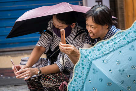 Yangshuo, China - August 2019 : Two older Chinese women, one of which has face covered in a protective mask and holding umbrella,  playing with their smartphones on a street in townのeditorial素材