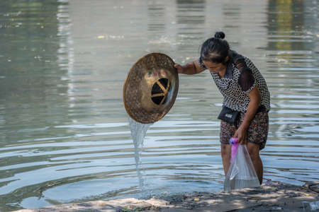 Feng Huang, China -  August 2019 : Old woman standing in shallow water on the riverbank, holding plastic bag with water and washing her conical wicker hat to chill out on a hot summer dayのeditorial素材