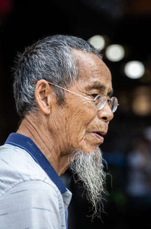 Feng Huang, China -  August 2019 : Portrait of an old chinese man with a grey beard wearing small...