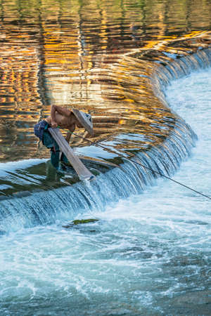 Feng Huang, China -  August 2019 : Fisherman with a traditional triangular chinese hat standing in waters of Tuo river flowing through the centre of Feng huang Old Town and catching fishesのeditorial素材