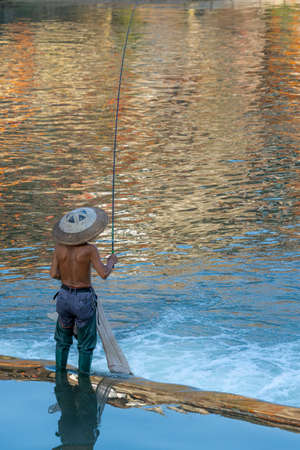 Feng Huang, China -  August 2019 : Fisherman with a traditional triangular chinese hat standing in waters of Tuo river flowing through the centre of Feng huang Old Town and catching fishesのeditorial素材