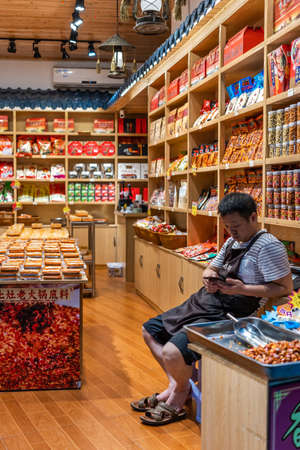 Chongqing, China -  August 2019 : Bored chinese male seller checking his smartphone while sitting and waiting for customers in his snack and food shop, Ci Qi Kou Old townのeditorial素材