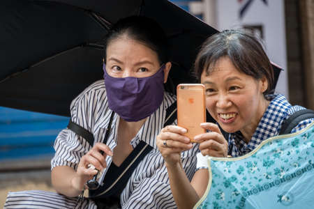 Yangshuo, China - August 2019 : Two older Chinese women, one of which has face covered in a protective mask and holding umbrella,  taking selfie with a smartphone on a street in townのeditorial素材