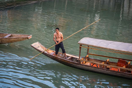 Feng Huang, China -  August 2019 : Half naked bare chest Chinese boatman with a long pole used for steering and pushing, standing on old wooden boat on the Tuo riverのeditorial素材