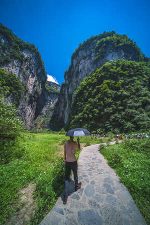 Wulong, China - August 2019 : Tourist with an umbrella providing shelter and shade from the blazing sun walking on the narrow mountain trekking path in the gorge valley among stunning rock formations, Wulong National Parkのeditorial素材