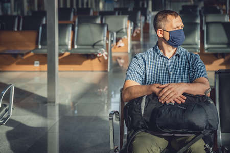 Middle aged male man with a backpack, wearing protective face mask, sitting alone in almost empty airport lounge waiting for the departure, traveling in the time of covid19 pandemicの写真素材