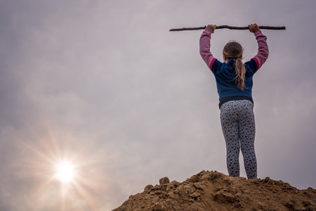 A low angle shot of a young kid holding up a wooden stick and standing on a hill
