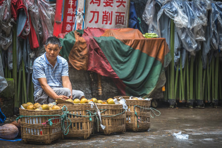 Zigong, China - July 2019 : Chinese man sitting on the pavement and selling vegetables stored in wicker bamboo baskets, Zigong town, Sichuan Provinceのeditorial素材